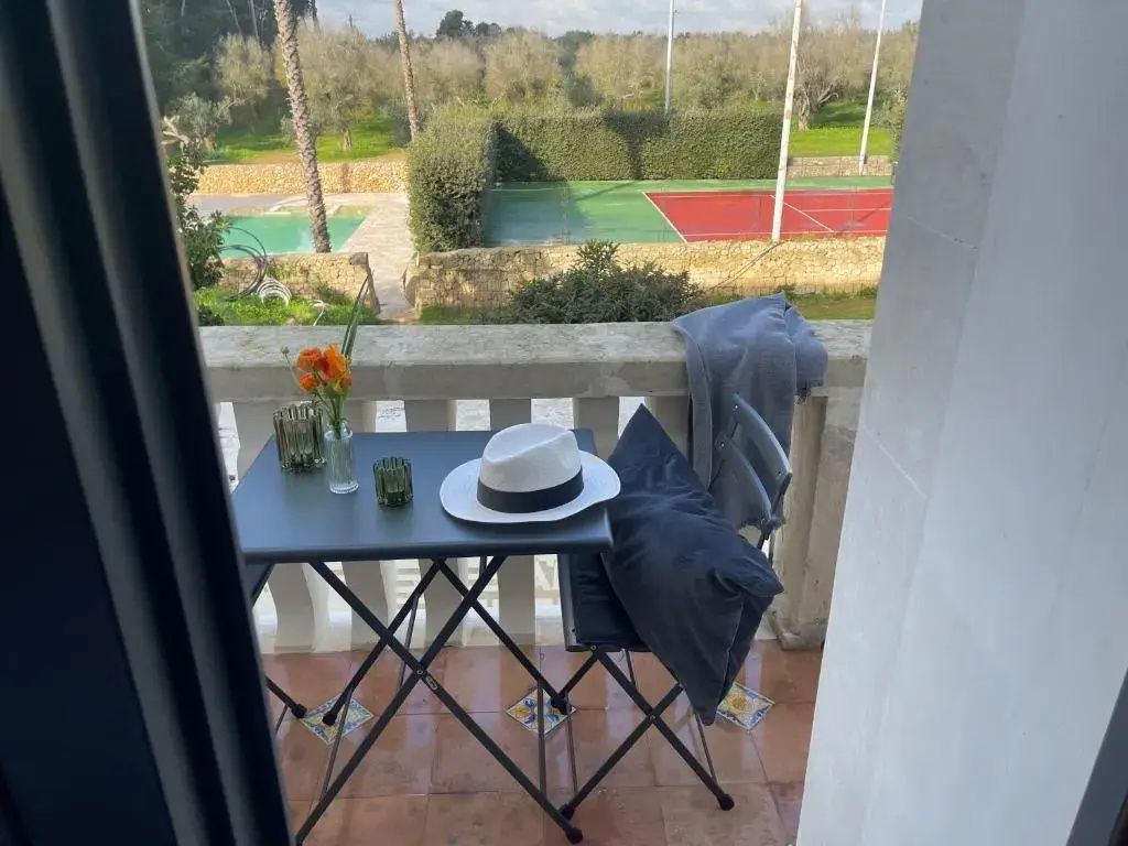 Blick von einem Balkon mit kleiner Sitzgruppe auf einen Tennisplatz und Olivenbäume.