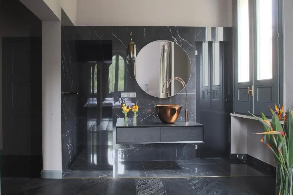 Badezimmer mit dunklem Marmor, einem kupferfarbenen Waschbecken und rundem Spiegel.