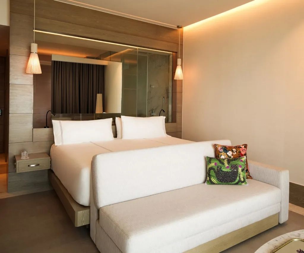 Doppelbett mit hellen Kissen und einer angrenzenden hellen Couch in einem hellen Hotelzimmer.