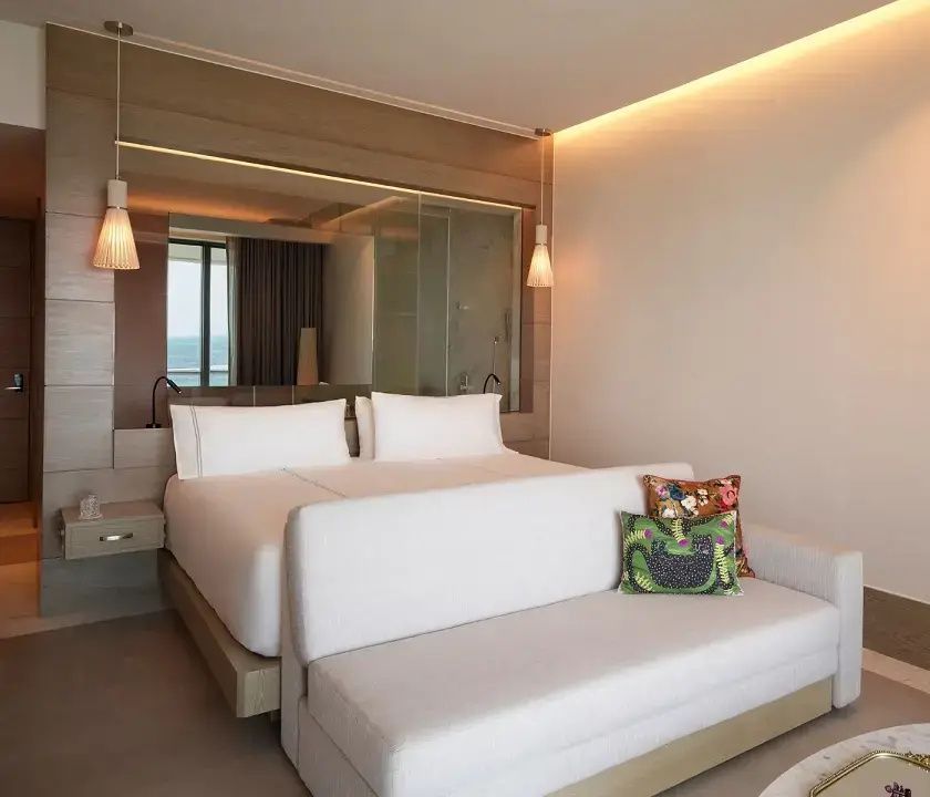 Doppelbett im hellen Hotelzimmer mit einem weißen Sofa davor und warmem Licht.