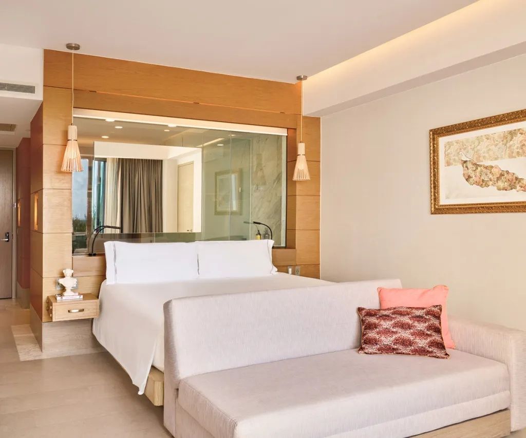 Helles Hotelzimmer mit Doppelbett, weißem Sofa davor und Glasfenster zum Badezimmer.