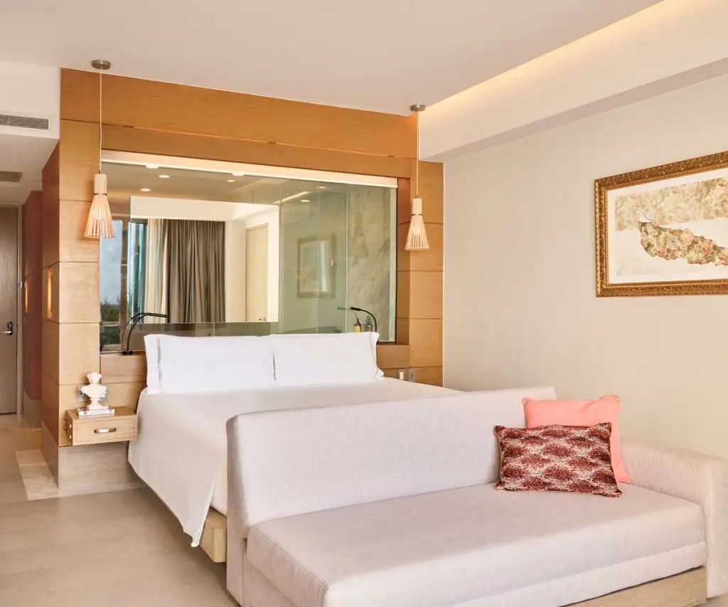 Hell eingerichtetes Hotelzimmer mit Doppelbett, hellem Sofabett und Glaswand zum Badezimmer.