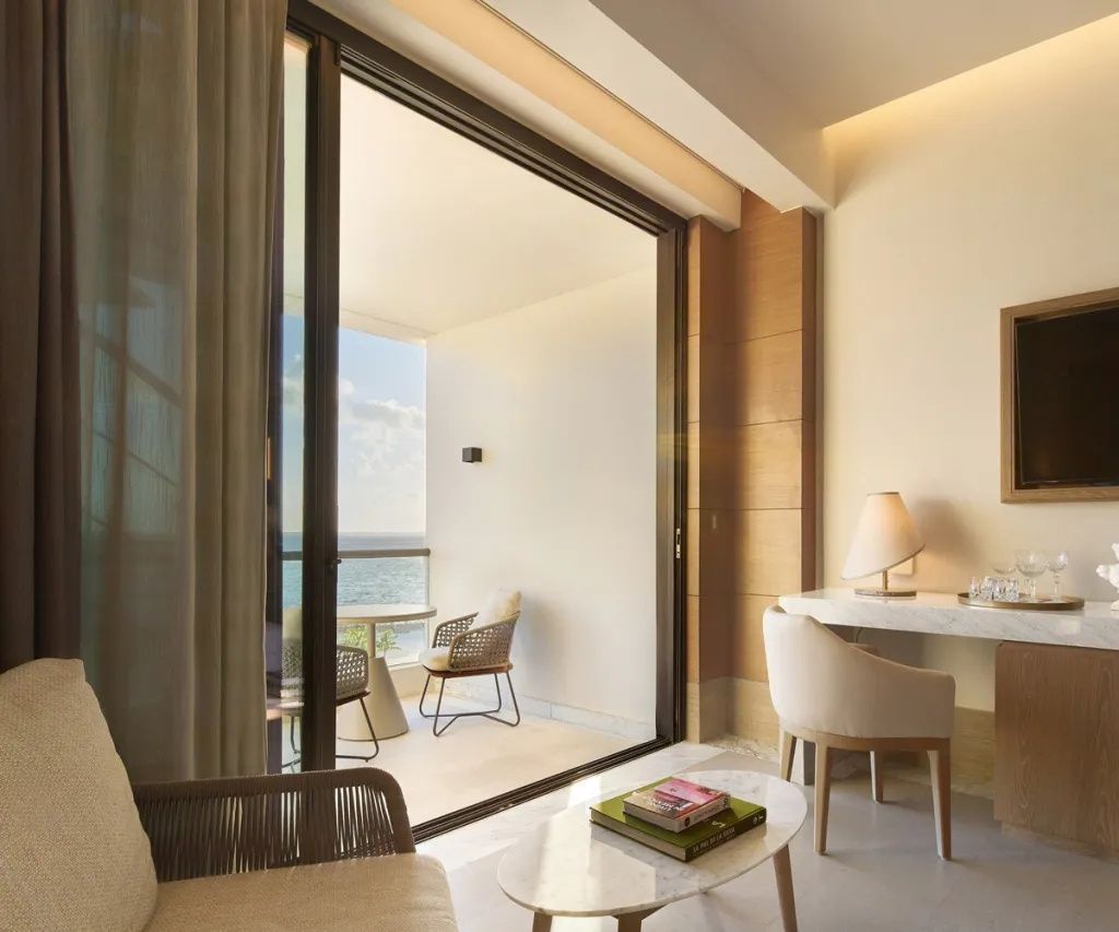 Hotelzimmer mit Schreibtisch, Sitzbereich und gläserner Schiebetür zum Balkon mit Meerblick.