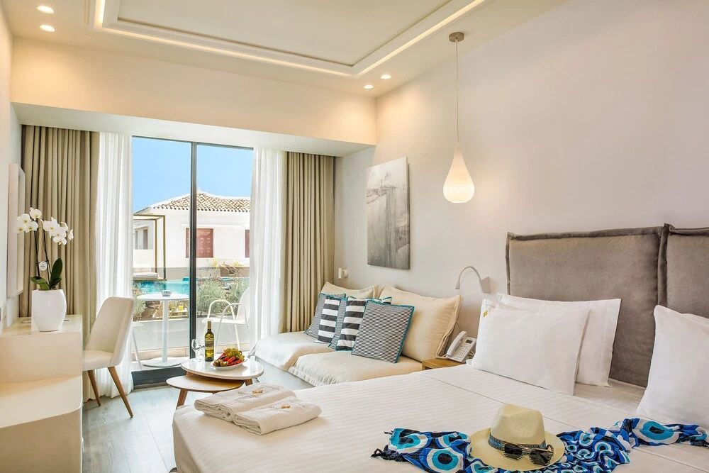 Hell eingerichtetes Hotelzimmer mit Doppelbett, Sitzbereich und Blick auf die Terrasse mit Pool.