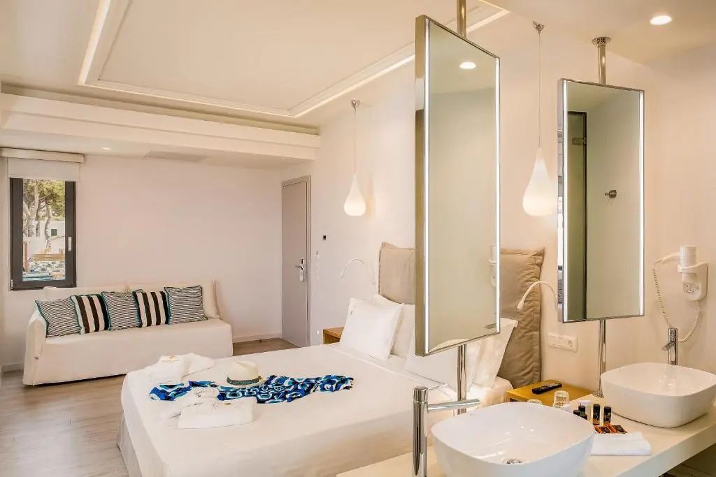 Modernes Hotelzimmer mit weißem Doppelbett, Waschtisch im Vordergrund und Sitzecke.