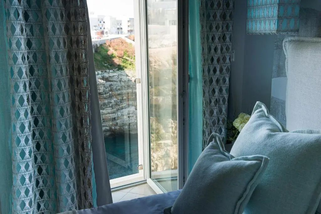 Blick durch ein geöffnetes Fenster auf eine felsige Meeresbucht mit türkisfarbenem Wasser.