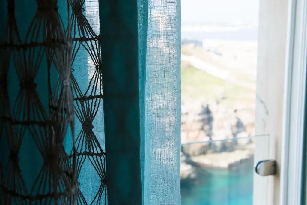 Blick durch einen blauen Vorhang auf den Balkon und das dahinterliegende Meer.