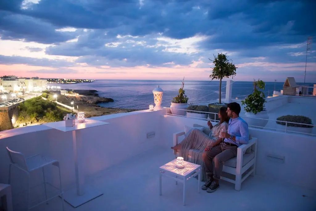 Terrasse bei Dämmerung mit Blick auf das Meer und die Küste sowie sitzendem Paar auf weißen Möbeln.