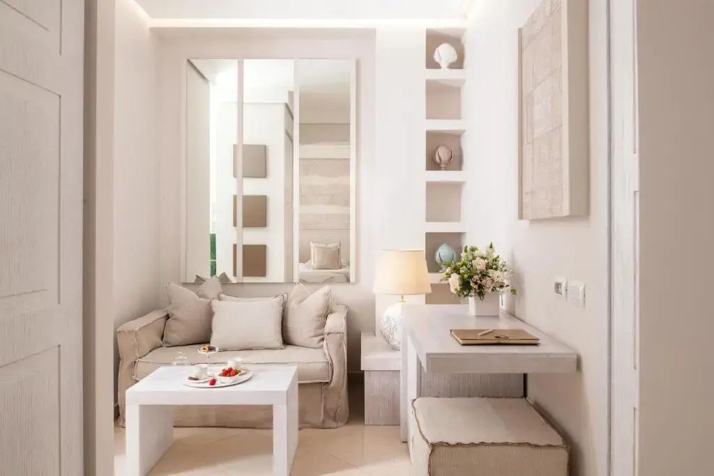 Sitzecke in einer Suite mit hellem Sofa, weißem Tisch, Schreibtisch und Wandregal.