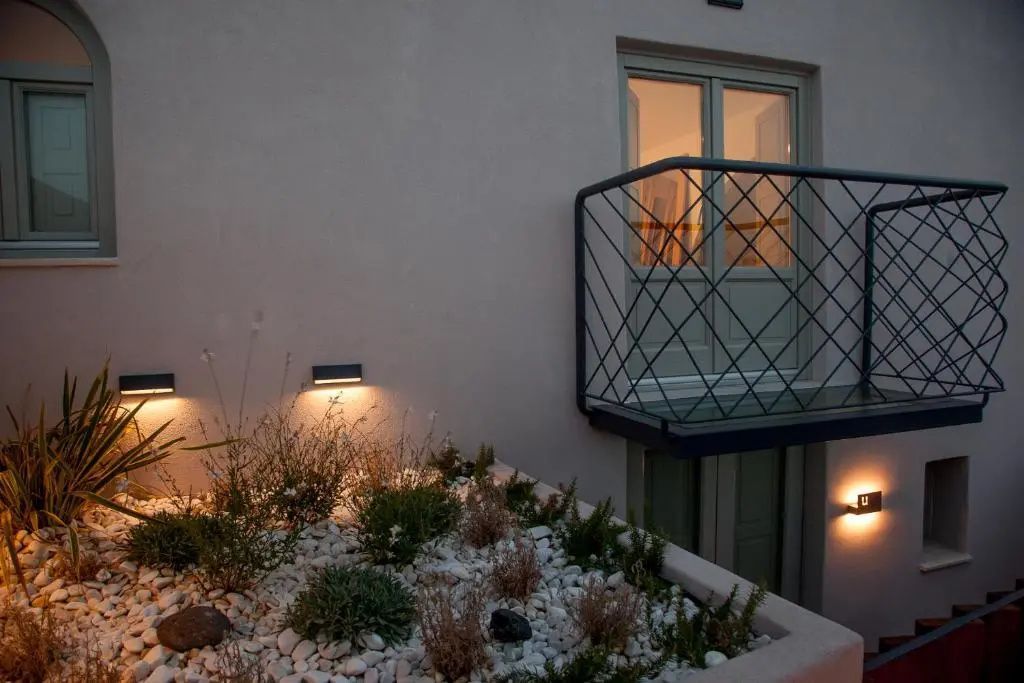 Beleuchtete Außenwand mit bepflanztem Beet und einem kleinen Balkon bei Dämmerung.