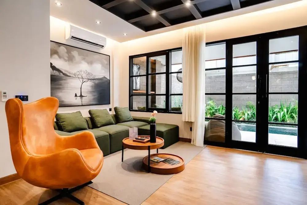 Wohnbereich mit grünem Ecksofa, orangefarbenem Sessel, Holztisch und großer Fensterfront.