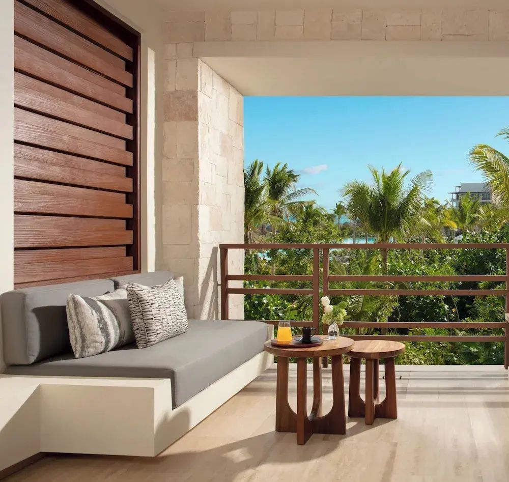 Überdachter Balkon mit grauem Sofa, Holztischen und Blick auf grüne Palmen.