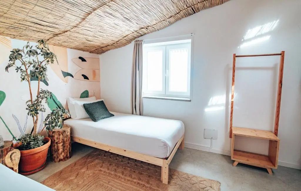 Einzelbett aus Holz in einem hellen Zimmer mit Strohdecke und Wandbemalung.