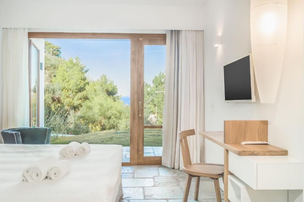 Helles Hotelzimmer mit Steinboden, Holzschreibtisch und Ausblick auf Bäume und das Meer.