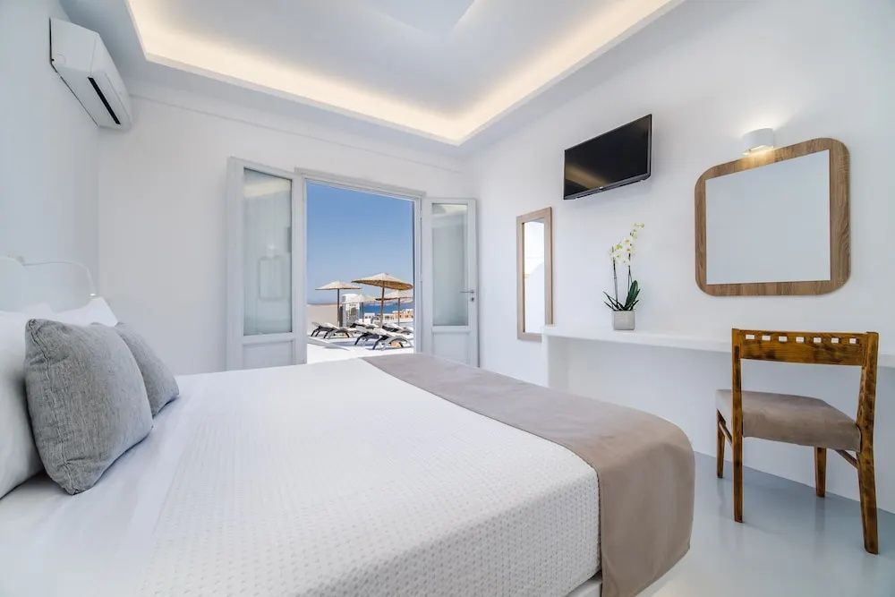 Weiß eingerichtetes Hotelzimmer mit Doppelbett, Fernseher und Blick durch die offene Balkontür nach draußen.