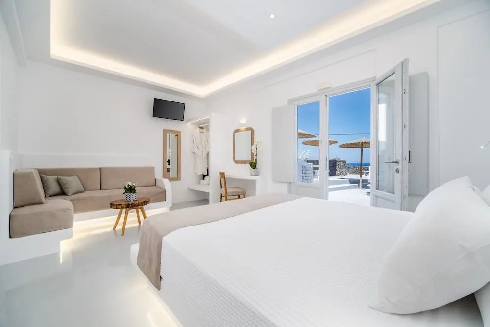 Geräumiges weißes Hotelzimmer mit Doppelbett, Beistelltisch, Sofa und offener Tür zur Terrasse.