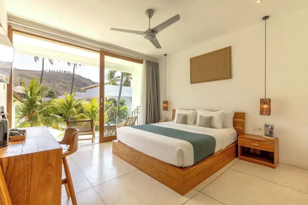 Helles Hotelzimmer mit großem Doppelbett, Deckenventilator und Schiebetür zum Balkon.