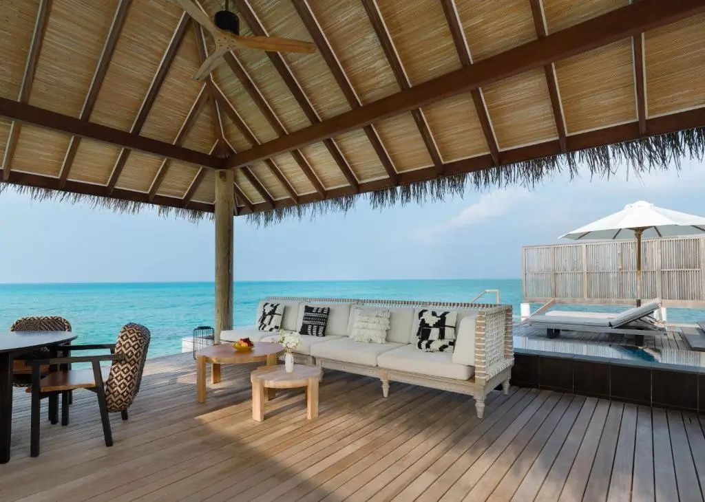 Überdachte Holzterrasse einer Wasservilla mit Sofa, Tischen und freiem Blick auf das türkisblau schimmernde Meer.