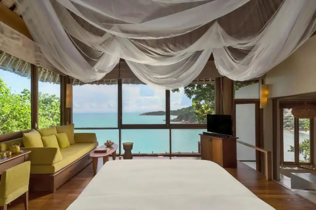Helles Schlafzimmer mit Bett unter Moskitonetz, gelber Couch und Panoramablick auf das türkisblaue Meer.