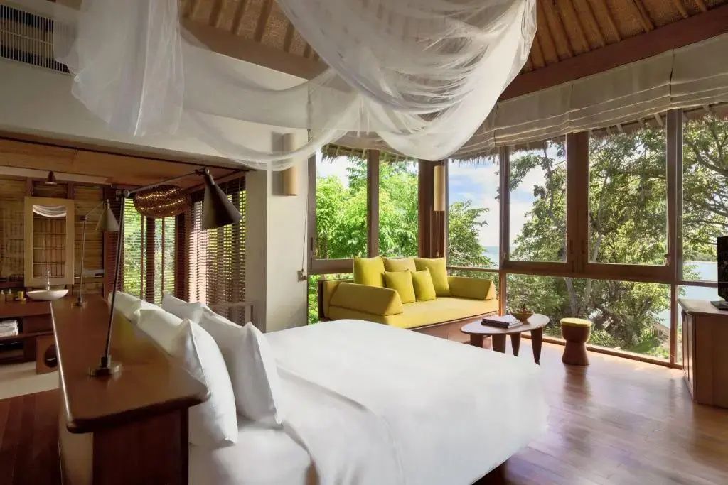 Helles Schlafzimmer einer Villa mit einem großen Doppelbett und gelbem Sofa an der Fensterfront.