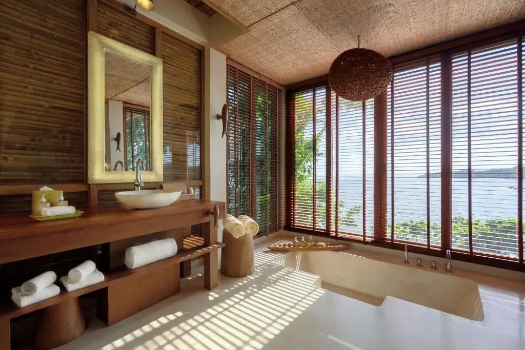 Badezimmer mit Holzwaschtisch, beleuchtetem Spiegel und Badewanne am Fenster mit Blick aufs Meer.
