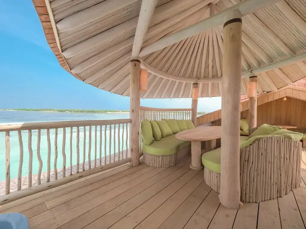 Überdachte Holzterrasse mit grüner Sitzecke und Blick auf das türkisfarbene Meer.