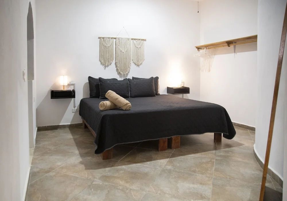 Doppelbett mit schwarzer Tagesdecke, braunen Handtüchern und Makramee an der weißen Wand.