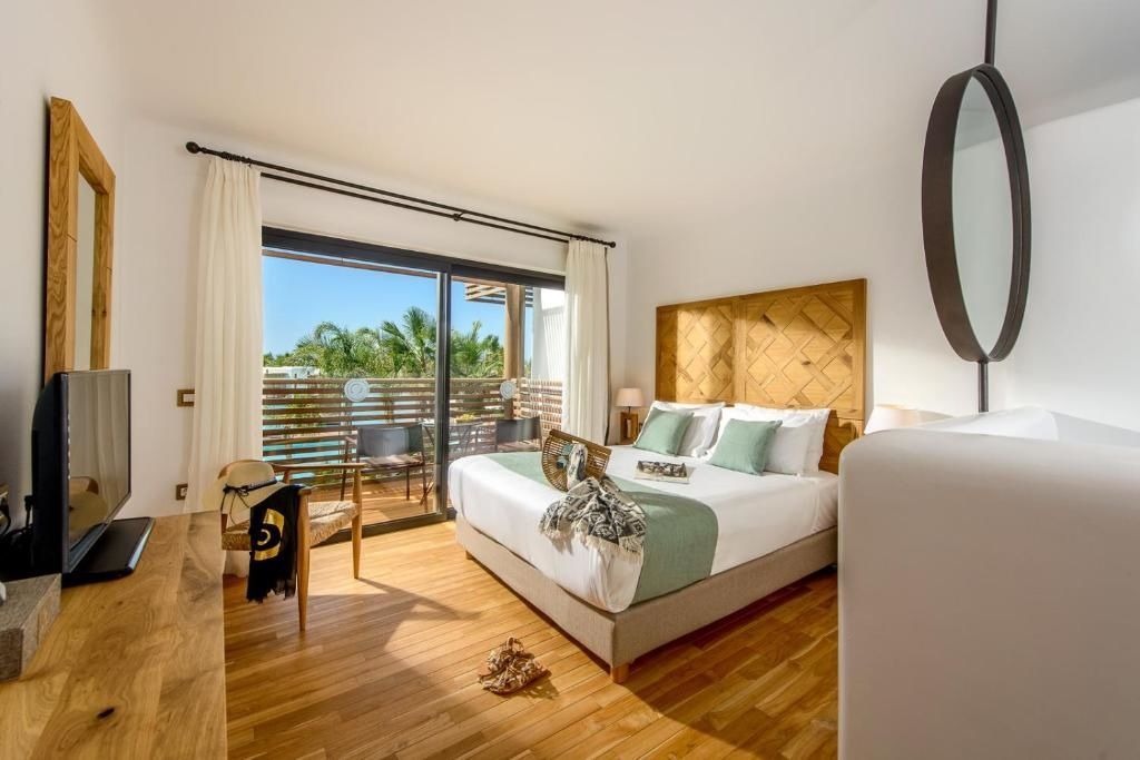Helles Hotelzimmer mit Doppelbett, Kopfteil aus Holz und Zugang zu einer Terrasse mit Sitzgelegenheiten.