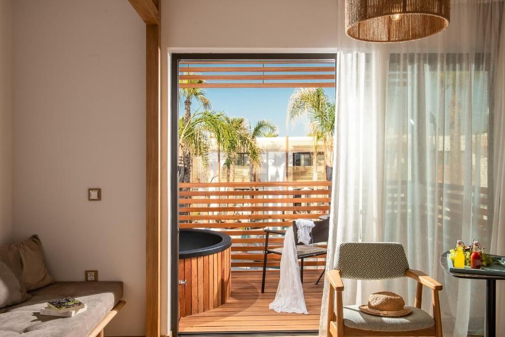Blick vom Zimmer auf den Balkon mit Whirlpool aus Holz, Stuhl und Palmen.