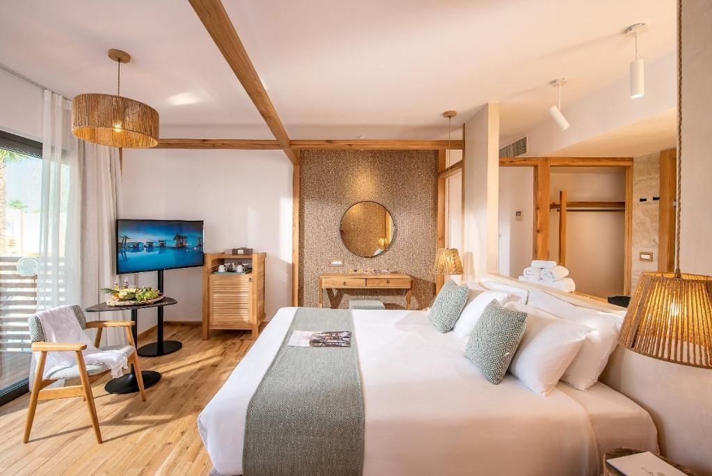 Doppelbett mit weißer Bettwäsche, Holzmöbeln und Fernseher im hellen Hotelzimmer.
