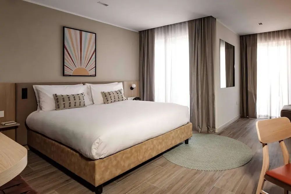Doppelbett mit weißer Bettwäsche, Wandbild und Fenster mit langen Vorhängen im Hotelzimmer.