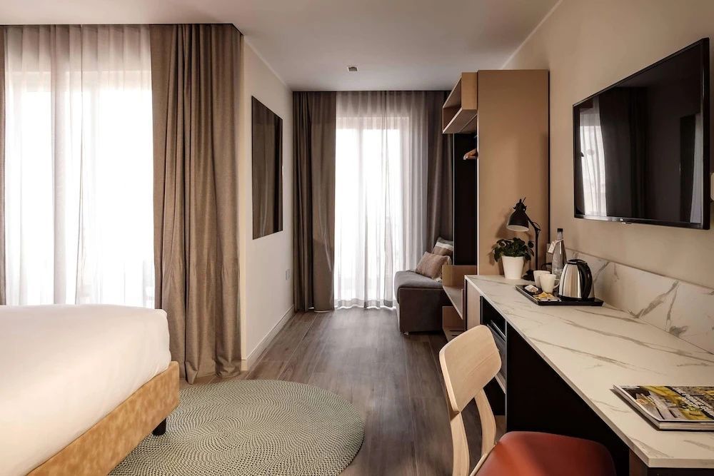 Sitzbereich mit Schreibtisch, Stuhl, Sofa und Fensterfront in einem hellen Hotelzimmer.