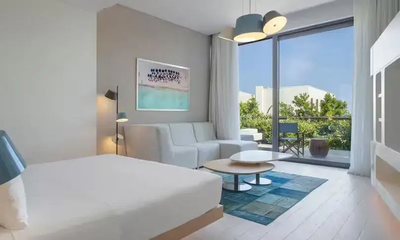 Helles Hotelzimmer mit beigen Möbeln, einem Sofa, blauem Teppich und Zugang zur Terrasse.