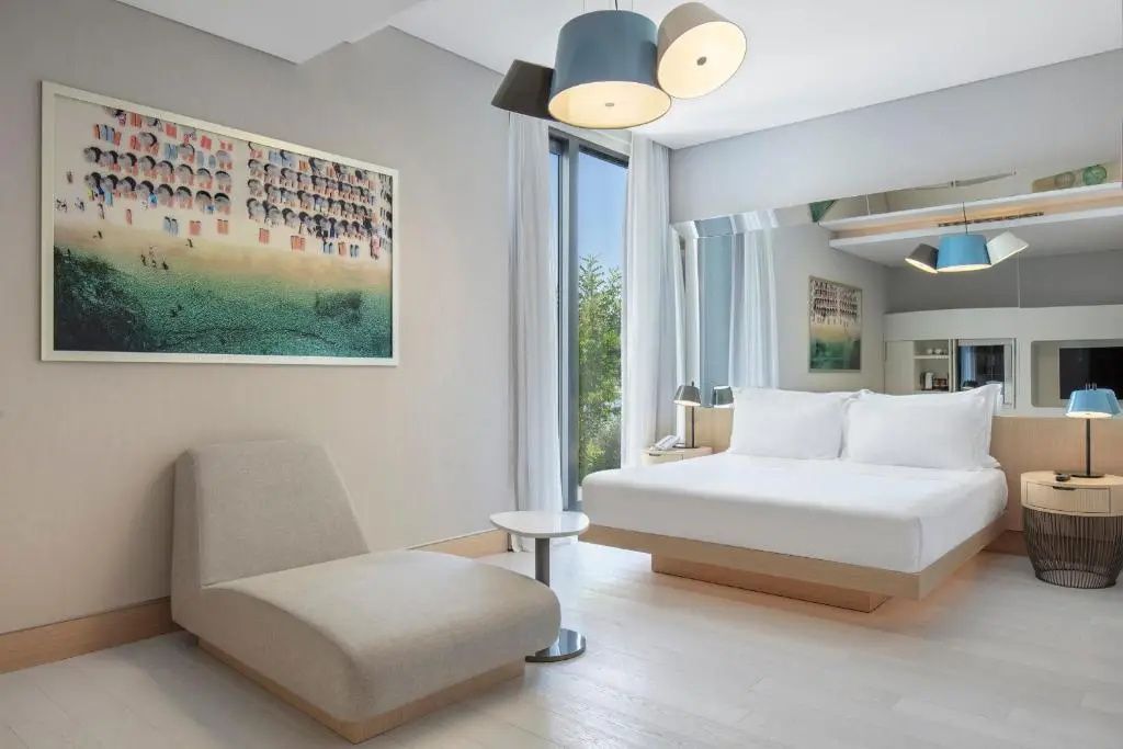 Helles Hotelzimmer mit Doppelbett, beigem Sessel und modernem Lampendesign an der Decke.