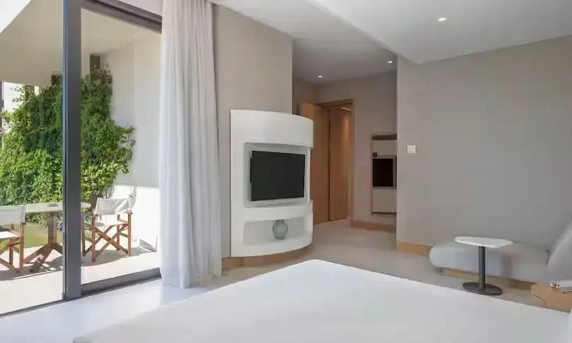 Helles Hotelzimmer mit weißem Bett, Wandfernseher und Blick auf die sonnige Terrasse.