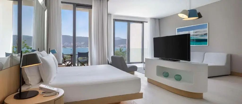 Doppelbett im hellen Hotelzimmer mit Fernseher und Meerblick durch bodentiefe Fenster.