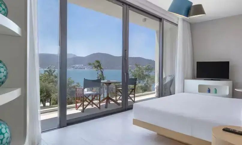Blick aus dem Hotelzimmer über den Balkon mit Sitzgelegenheiten auf das blaue Meer und Hügel.