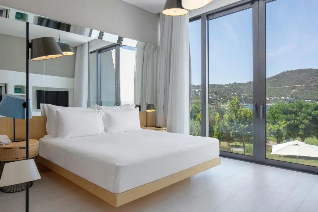 Helles Hotelzimmer mit großem Doppelbett und bodentiefen Fenstern mit Blick ins Grüne.