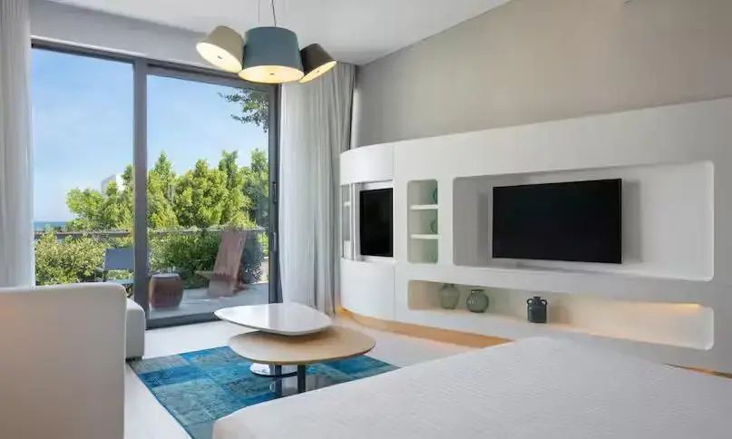 Modernes Hotelzimmer mit weißer Schrankwand, Fernsehern, blauem Teppich und großen Fenstern.