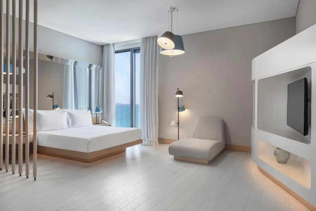 Schlafbereich mit weißem Bett, Sessel, modernen Hängelampen und Meerblick durch das Fenster.