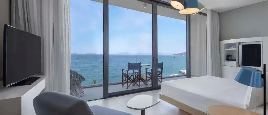 Blick durch große Glasfenster auf den Balkon mit Stühlen und das weitläufige Meer.