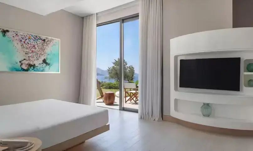 Schlafzimmer mit weißem Bett, Fernseher und großer Glas-Schiebetür mit Blick auf den Garten und das Meer.