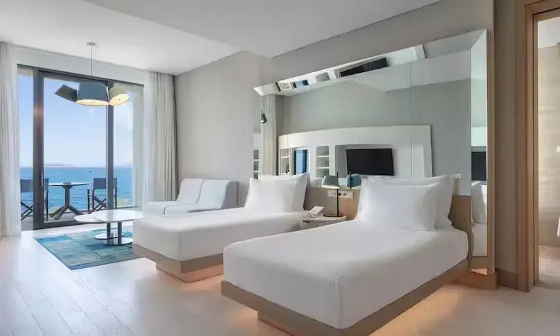 Modernes Hotelzimmer mit zwei Einzelbetten, spiegelnder Wand und Fensterfront mit Meerblick.