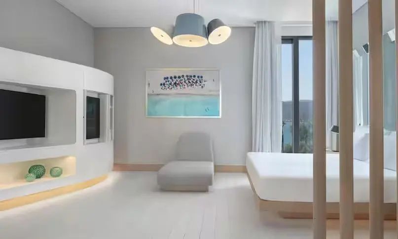 Zimmer mit weißem Bett, beigem Sessel, Kunstwerk an der Wand und Blick ins Freie.