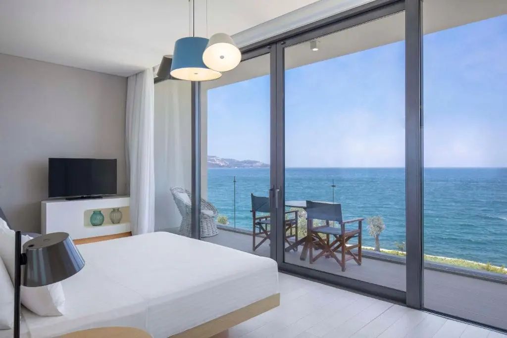 Helles Hotelzimmer mit Doppelbett und großem Panoramafenster zum Meer sowie Balkon.