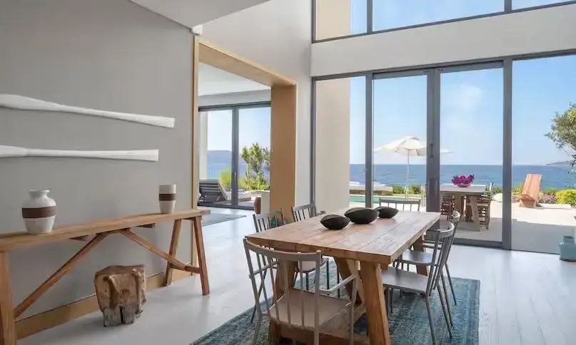 Essbereich mit Holz-Esstisch und Stühlen sowie bodentiefen Fenstern mit Blick auf Terrasse und Meer.