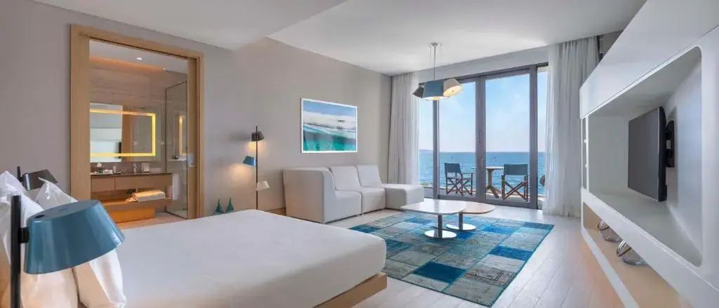 Bett und weiße Eckcouch auf blauem Teppich vor großem Balkonfenster mit Meerblick.