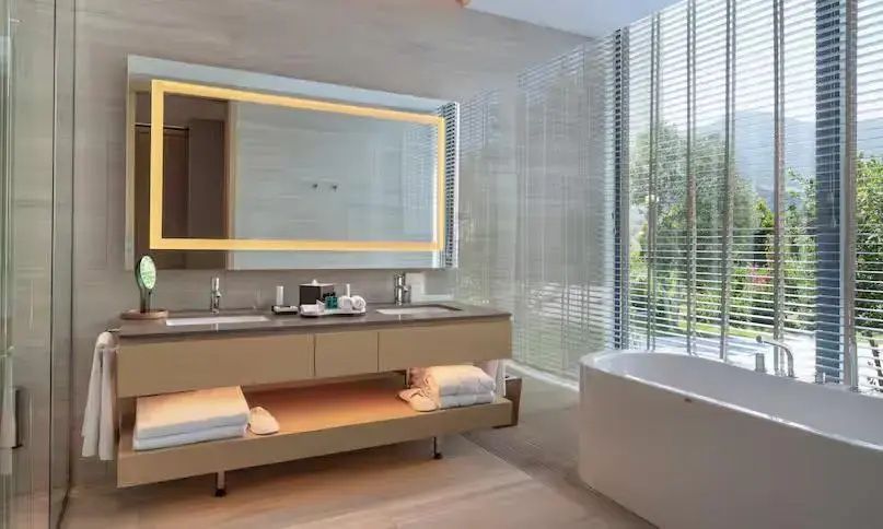 Badezimmer mit Doppelwaschtisch, beleuchtetem Spiegel und einer freistehenden Badewanne am Fenster.