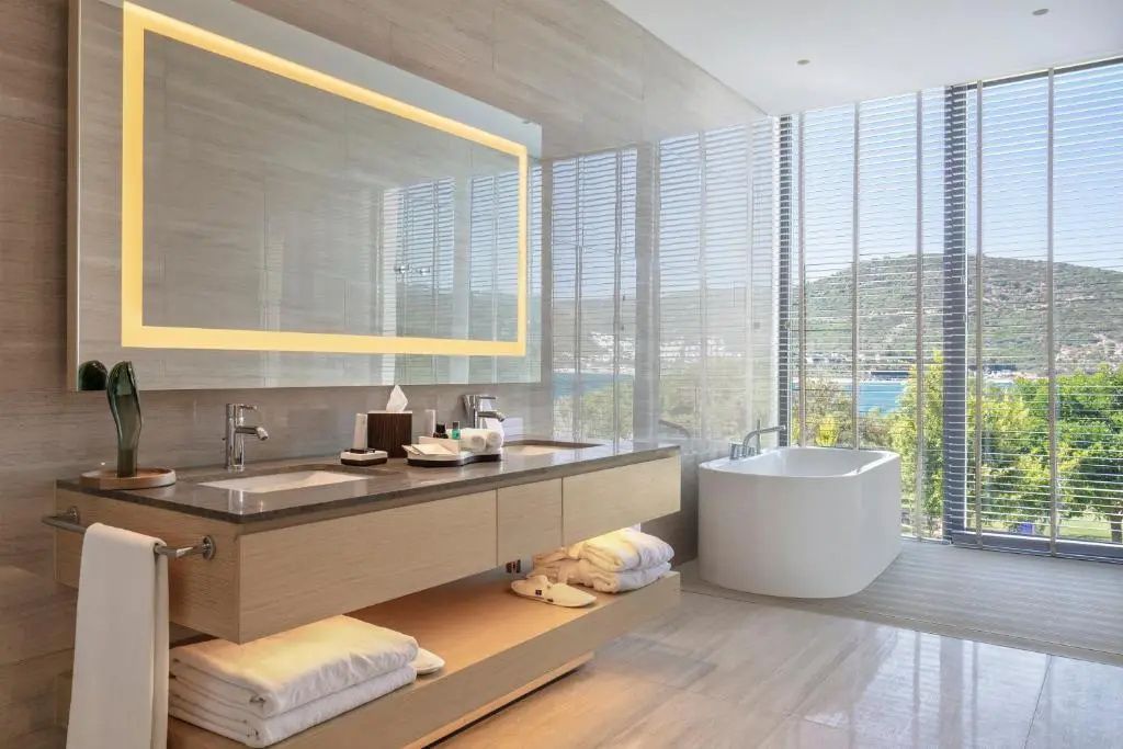 Badezimmer mit Doppelwaschtisch, beleuchtetem Spiegel und freistehender Badewanne am Fenster.