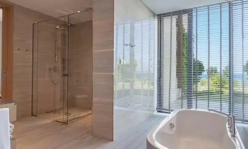 Badezimmer mit bodengleicher Glasdusche, Badewanne und großem Fenster mit Jalousie.